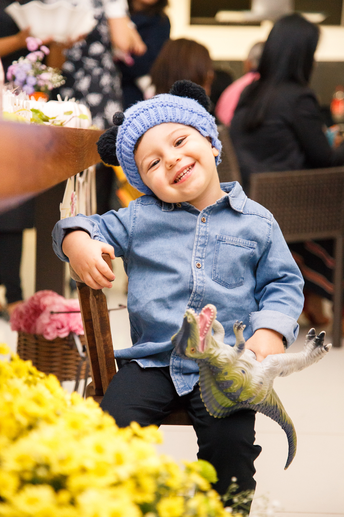 Criança com um gorro de bichinho sentado na cadeirinha em frente à mesa do bolo olhando para a câmera e sorrindo.