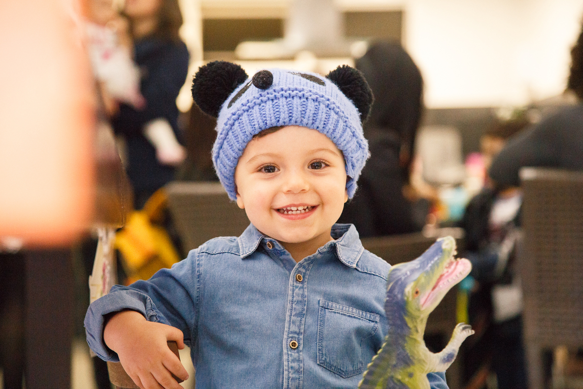Criança usando gorrinho de bichinho sentado em uma cadeirinha segurando um dinossauro, olhando para  foto e sorrindo.