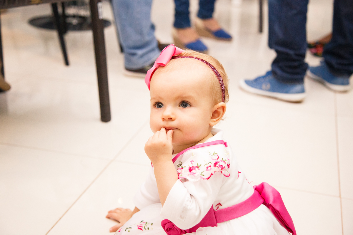Bebê aniversariante sentada no chão colocando um pedaço de comida na boca e prestando atenção em algo que acontece ao redor.