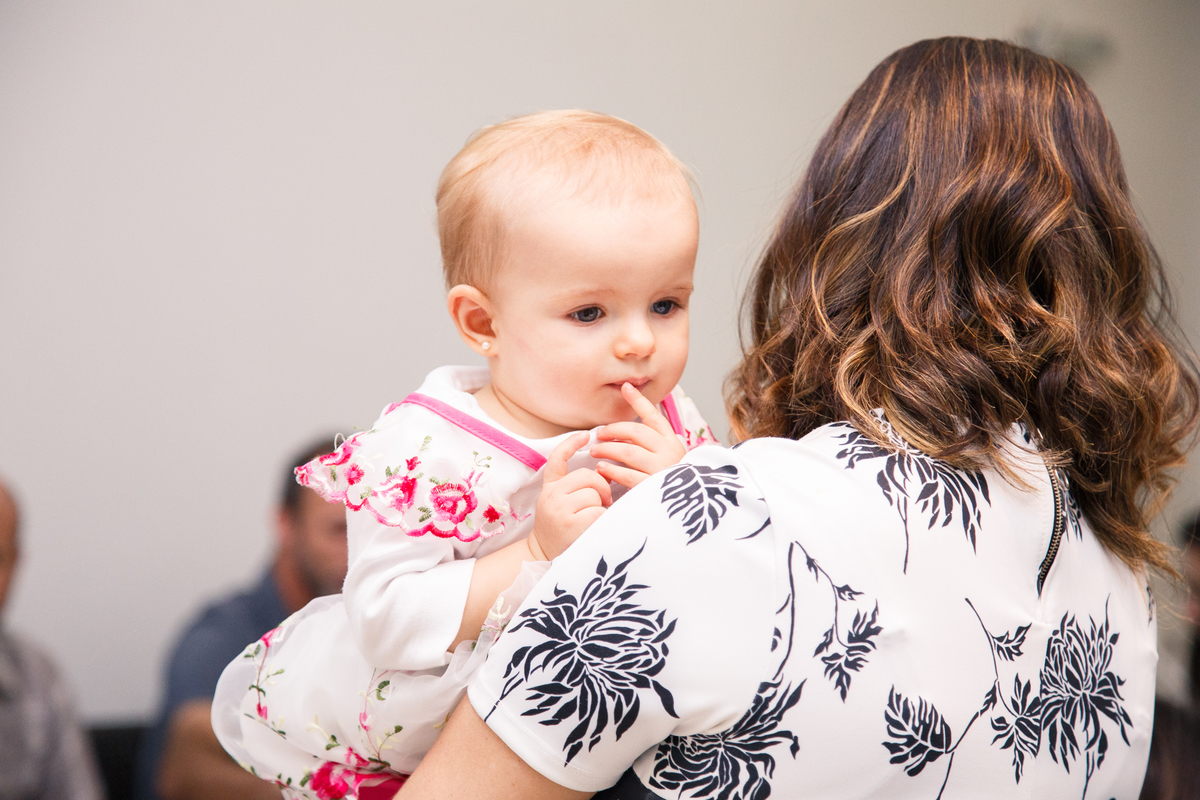 Criança no colo, olhando por sobre o ombro da mãe com uma expressão pensativa, um dedinho sobre o lábio.