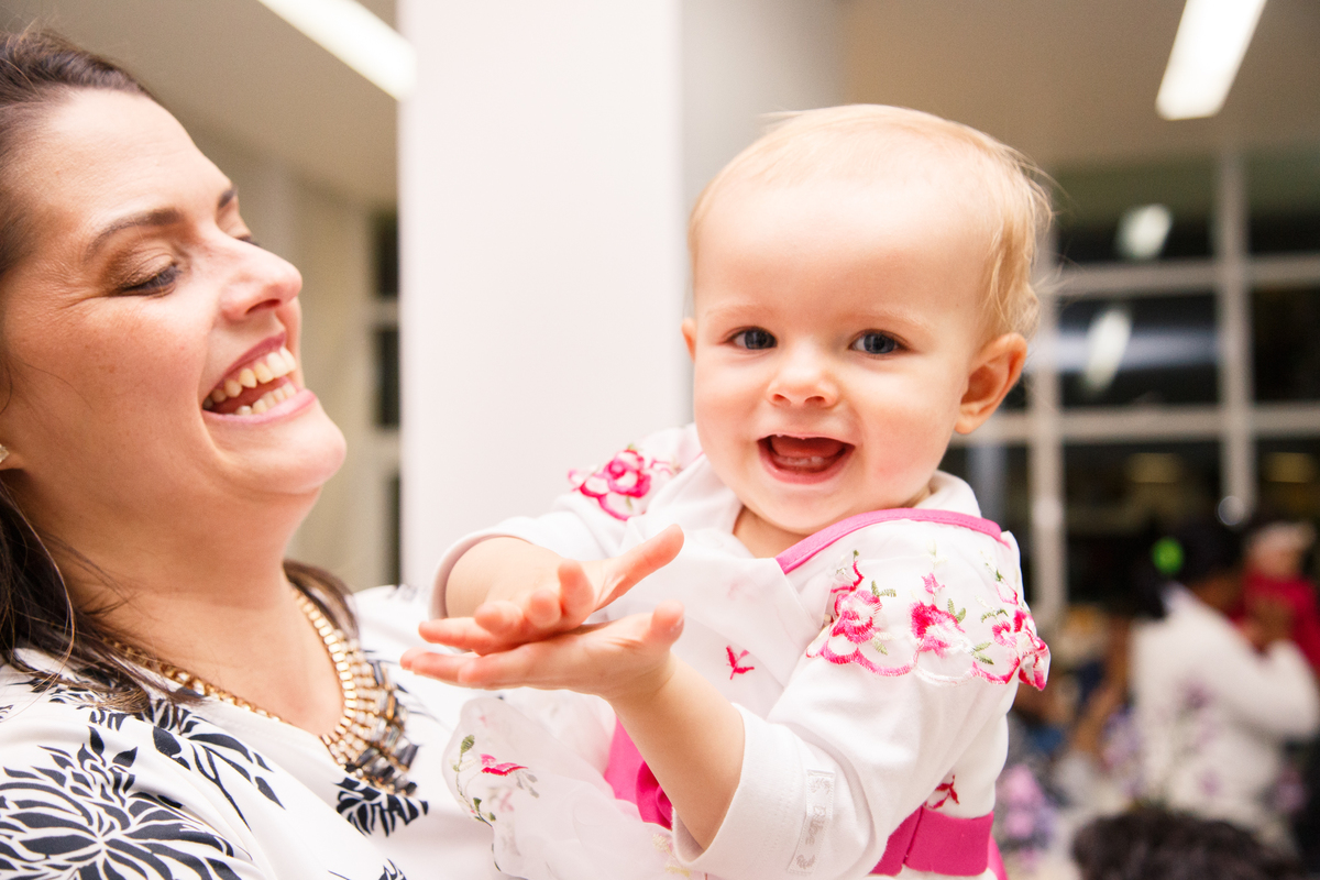 Bebê sorrindo no colo de sua mãe, batendo palminhas e olhando para a câmera.