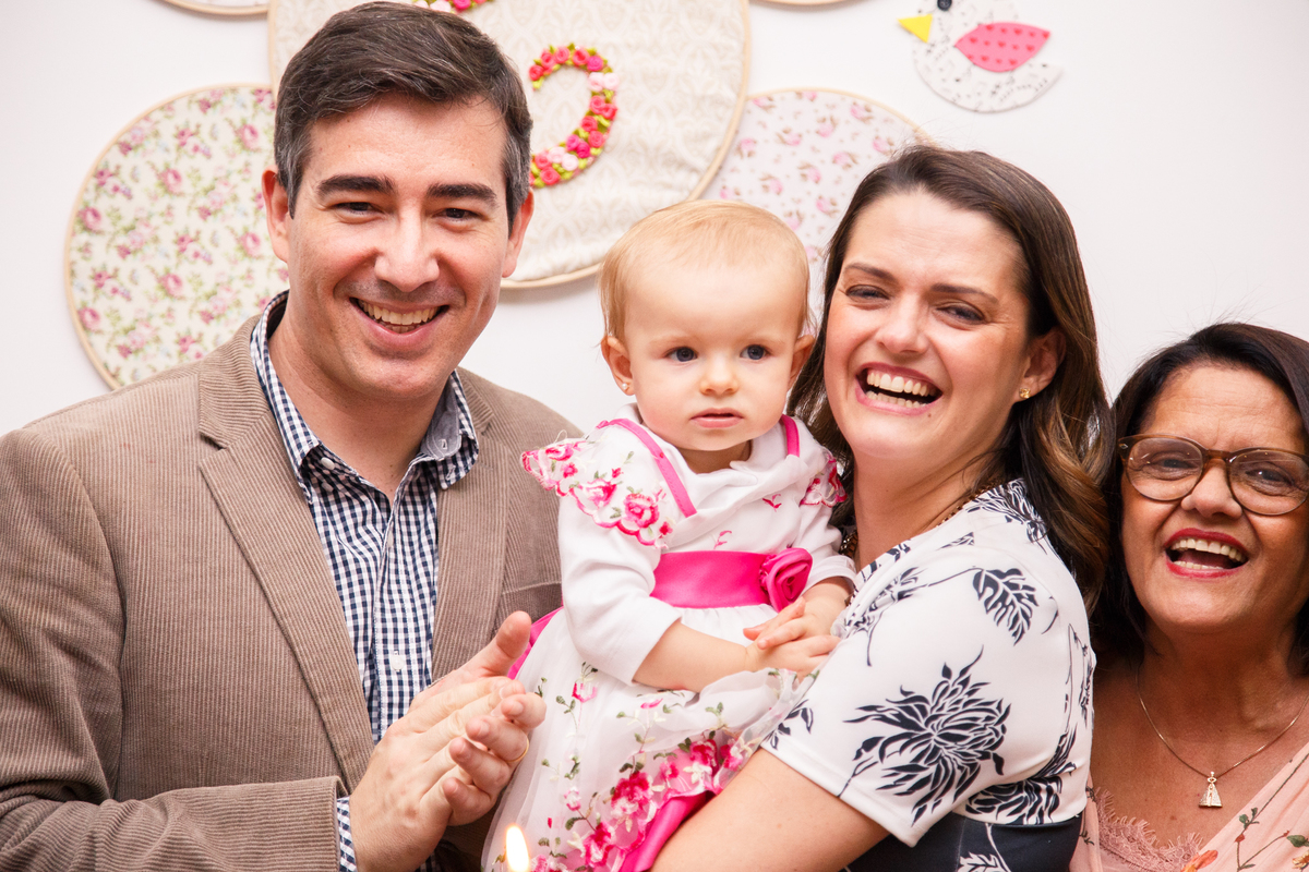 Pais com a bebê no colo atrás da mesa do bolo durante parabéns, todos olhando para a câmera e sorrindo.