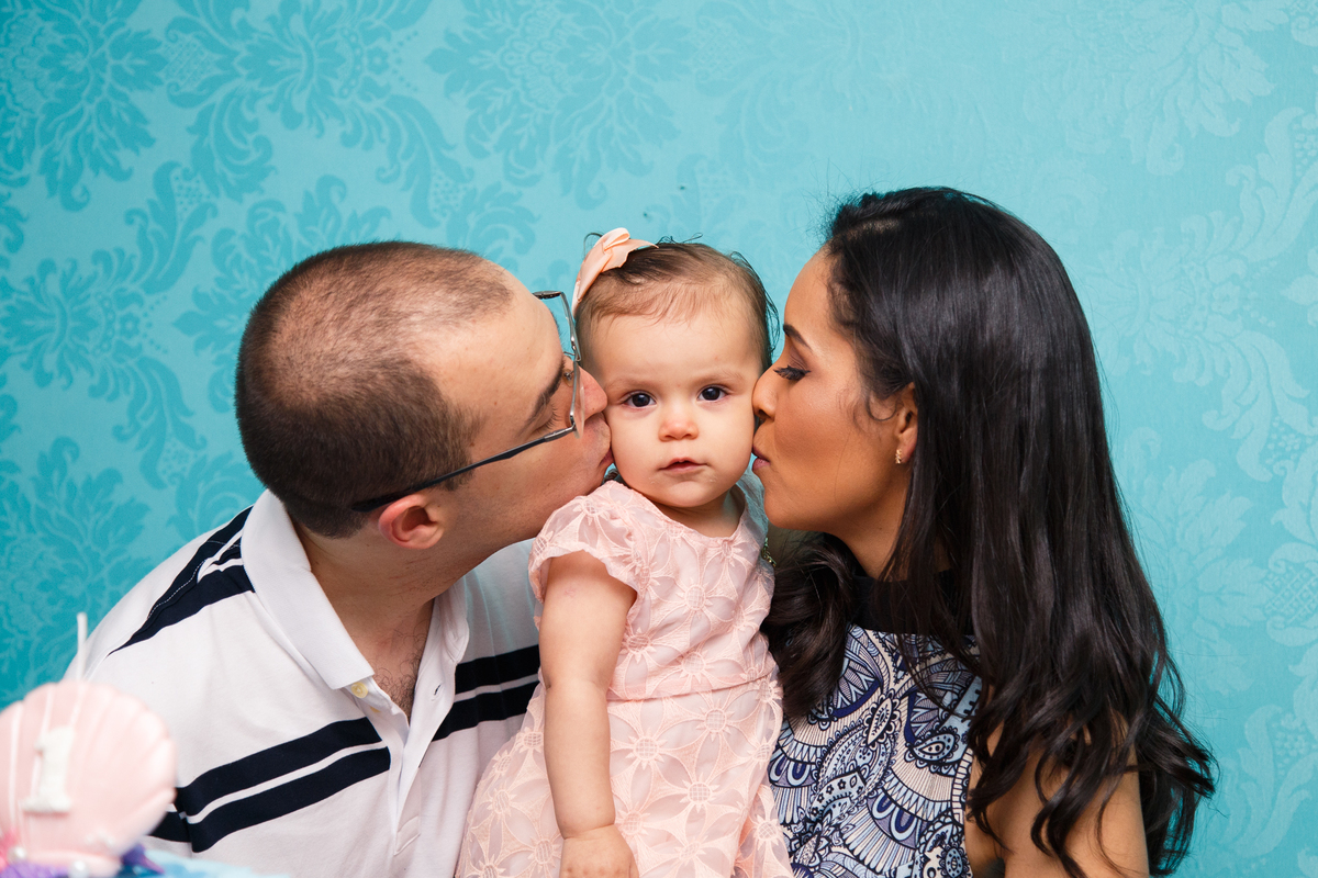 Bebê de um ano no colo dos pais ganhando um beijo na bochecha de cada um, um de cada lado.