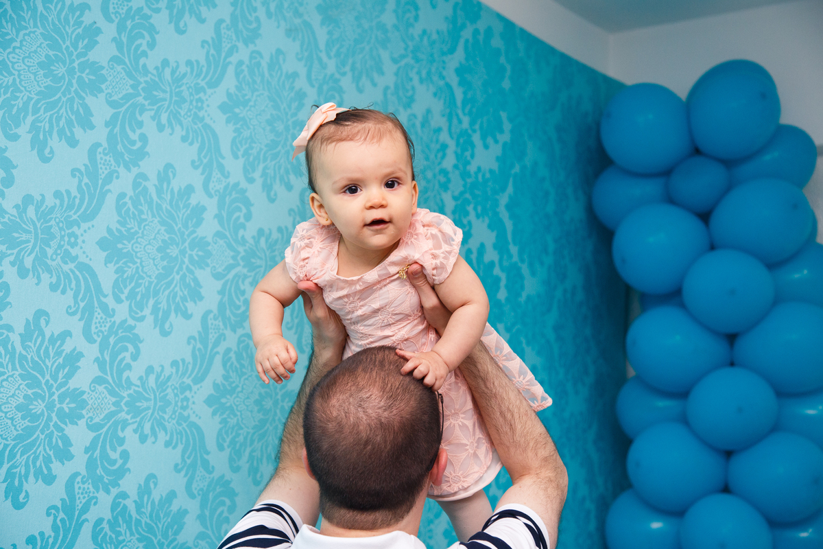 Pai brincando de levantar sua bebê no aniversário de 1 ano dela.O pai de costas e a bebê de frente olhando para a câmera.