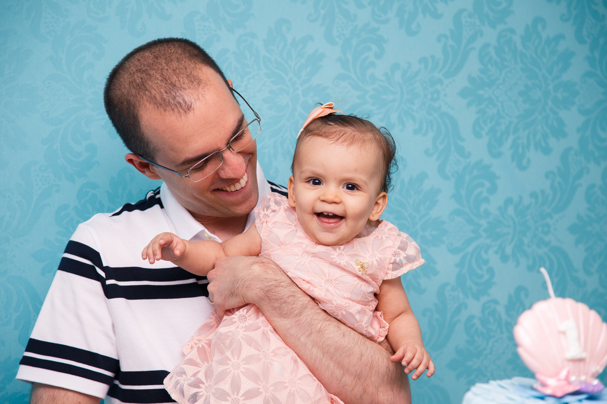 Bebê sorrindo no colo de seu pai. Ele olha para ela e ela olha para a câmera.