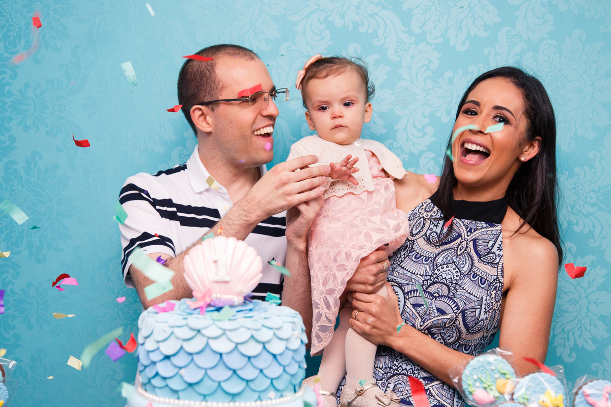 Pai e mãe com a bebê no colo comemorando o parabéns, com papéis picados caindo sobre eles.