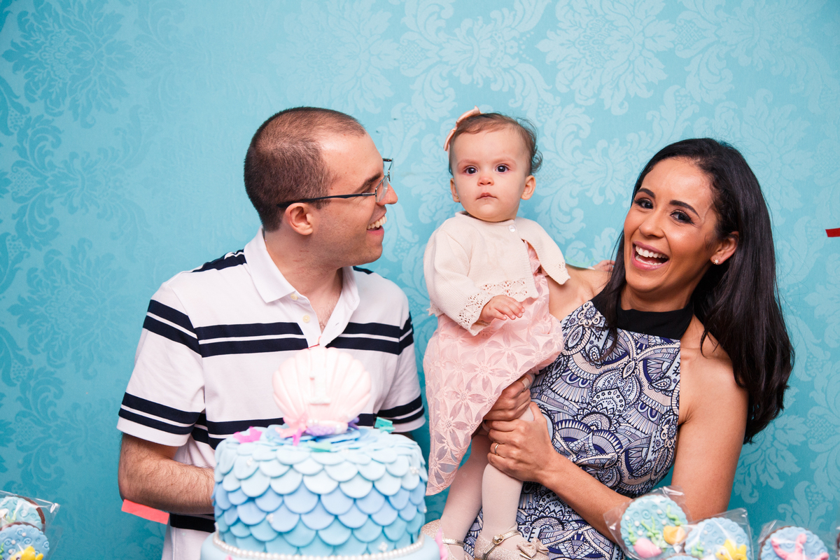 Mãe e pai com sua bebê no colo atrás da mesa do bolo durante os parabéns.