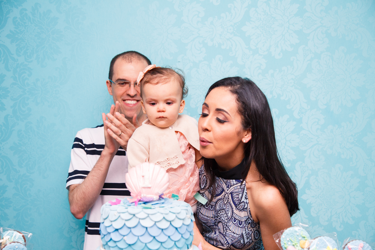 Mãe e pai com sua bebê no colo soprando a velinha de seu aniversário de 1 ano.