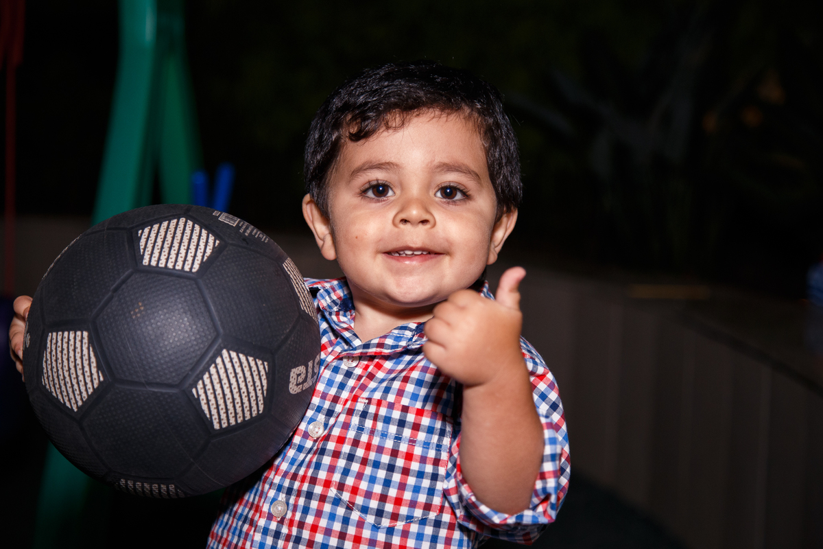 Criança segurando uma bola de futebol, olhando para a câmera e fazendo um joia com o polegar.