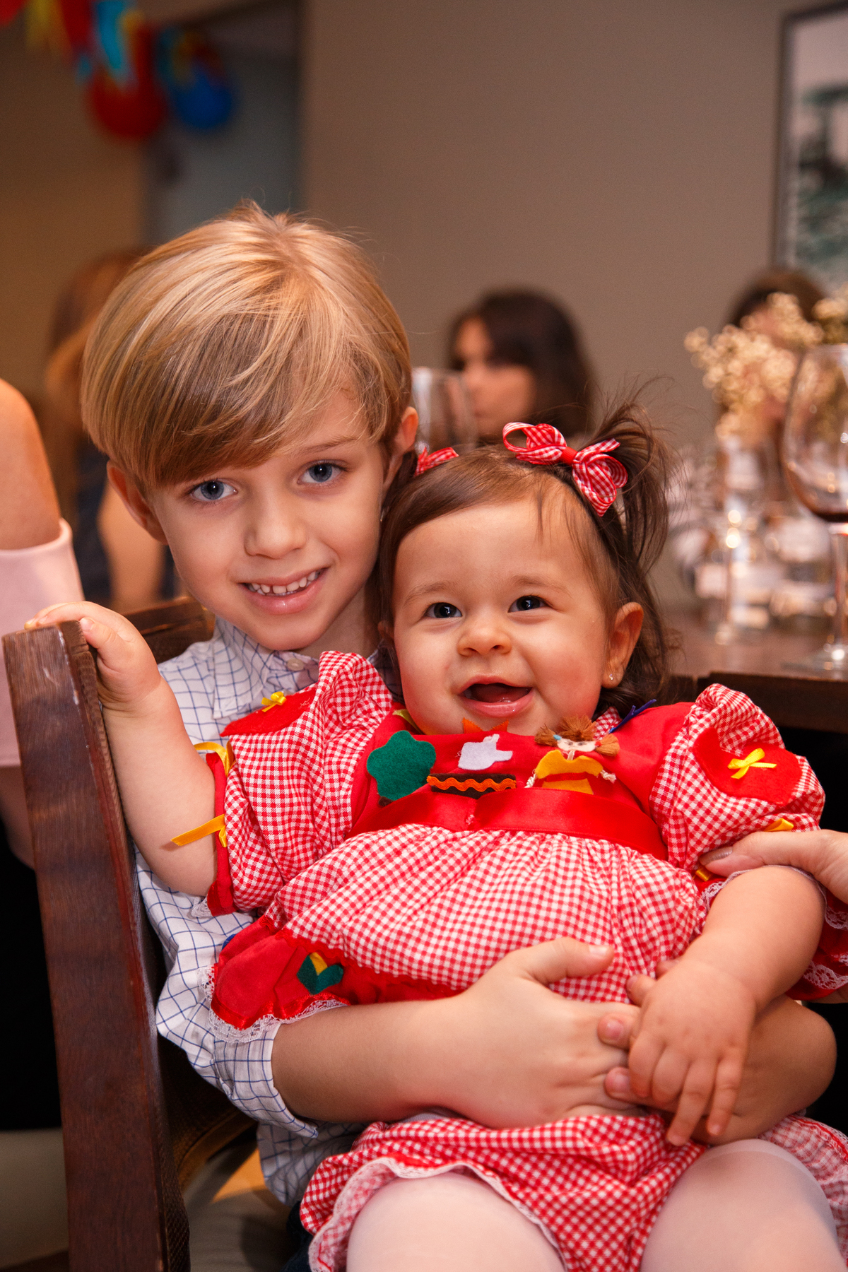 Criança segurando no colo a bebê aniversariante, ambos olhando para a câmera e sorrindo.