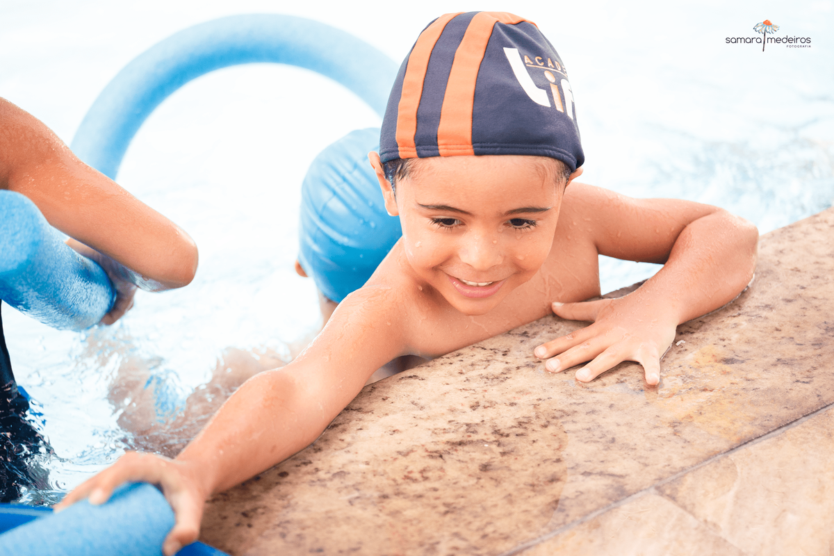 Criança na borda de uma piscina, do lado de dentro, tentando alcançar um espaguete que está do lado de fora.