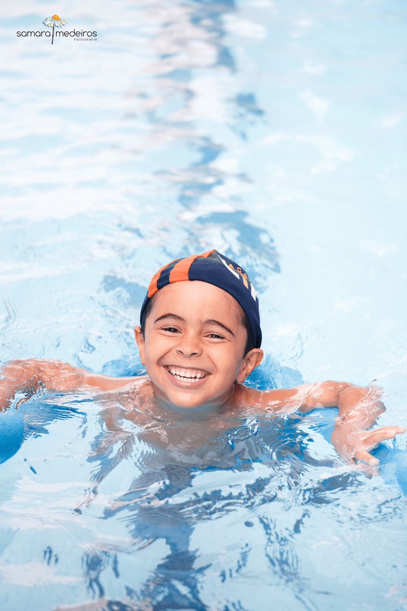 Criança dando uma gargalhada enquanto nada com ajuda de um espaguete durante aula de natação.