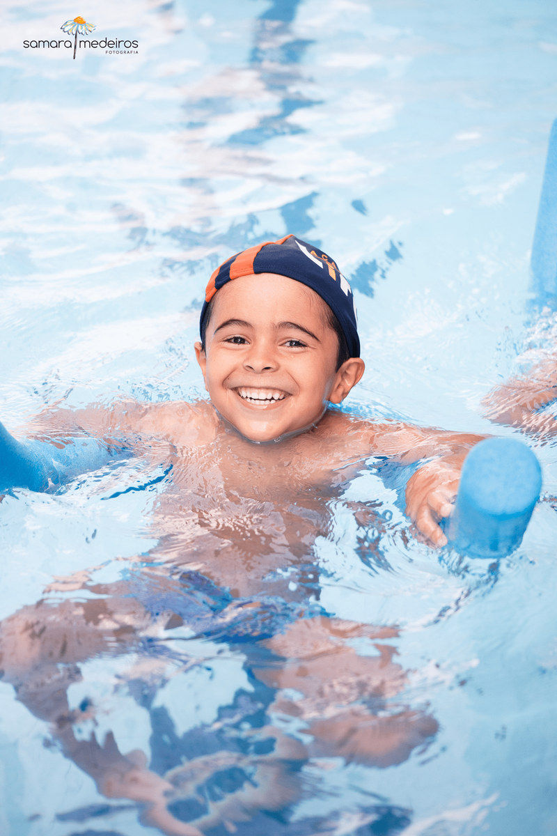 Criança com um enorme sorriso nadando em uma piscina com ajuda de um espaguete.