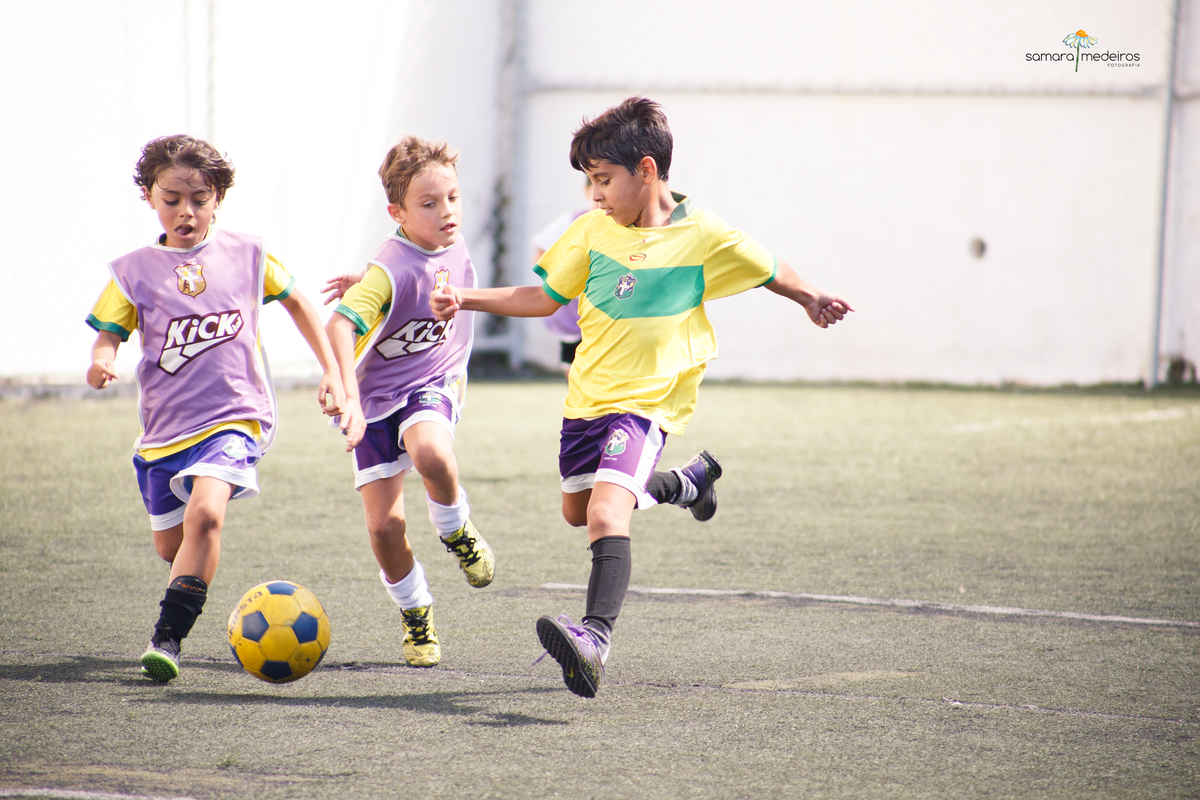 Três crianças correndo atrás de uma bola durante aula de futebol.