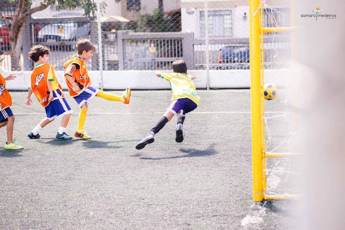 Crianças jogando futebol, uma chuta para o gol e a outra pula para tentar defender.