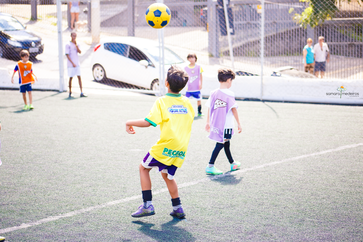 Crianças jogando futebol em aula, a bola está no ar e uma das crianças tenta mirar para dar uma cabeçada.