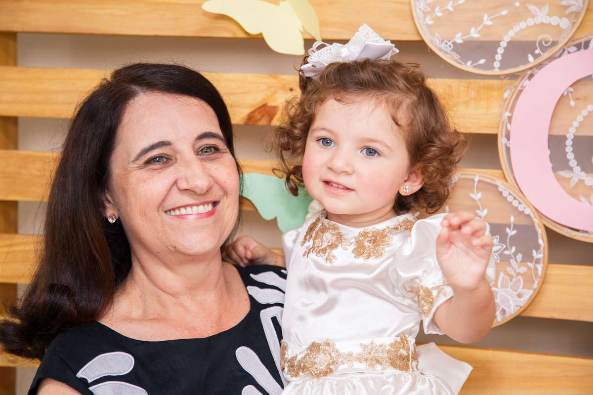 Criança aniversariante no colo de sua avó em frente ao painel da mesa do bolo.