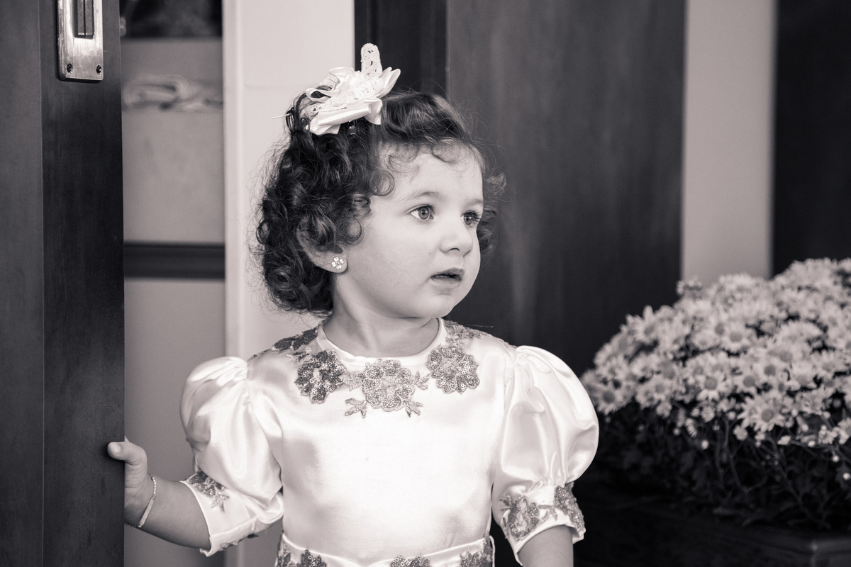 Foto em preto e branca de uma criança ao lado de um arranjo de flores observando as pessoas em sua festa de aniversário.