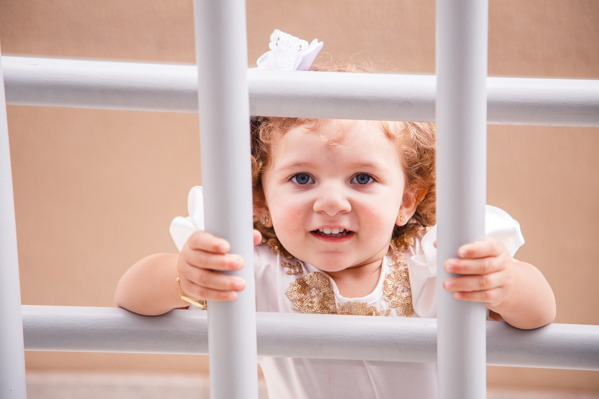 Criança atrás de barras brancas que compõem o corrimão da área de piscina da casa dela.