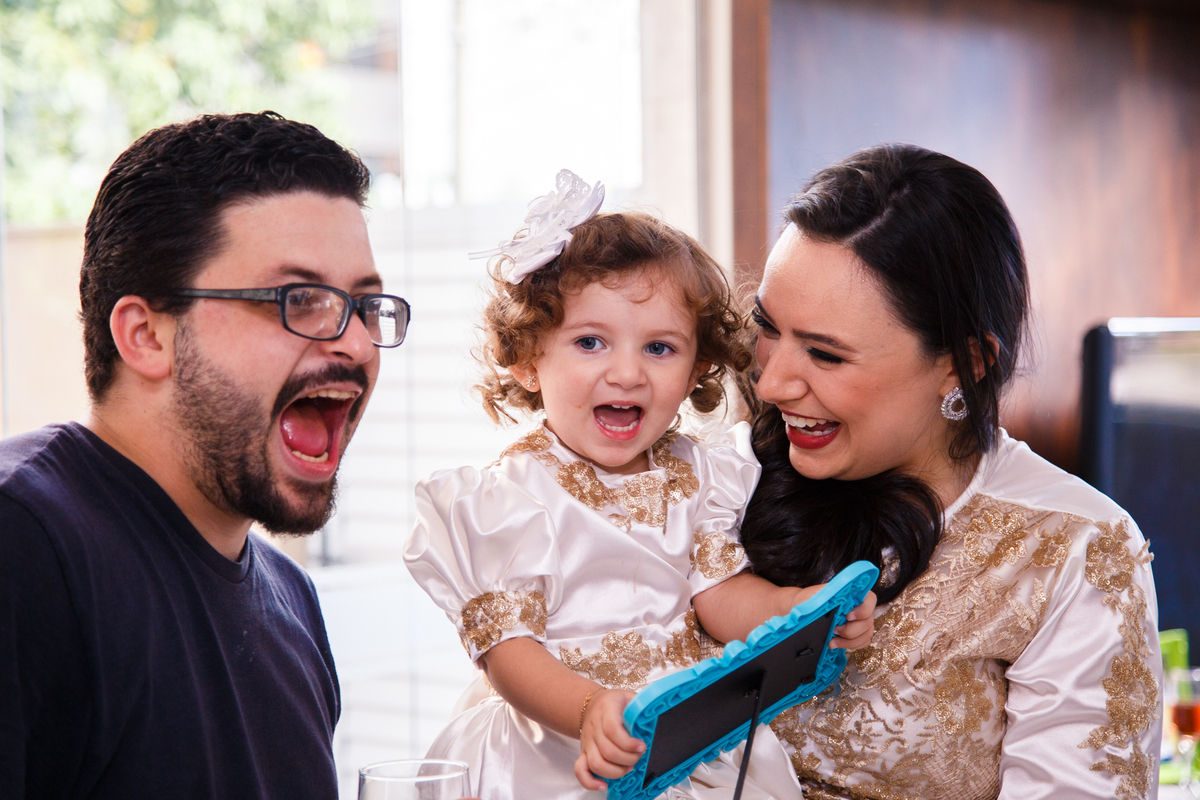 Criança no colo da mãe com um tio ao lado, os três brincando de fazer caretas, com a boca aberta.