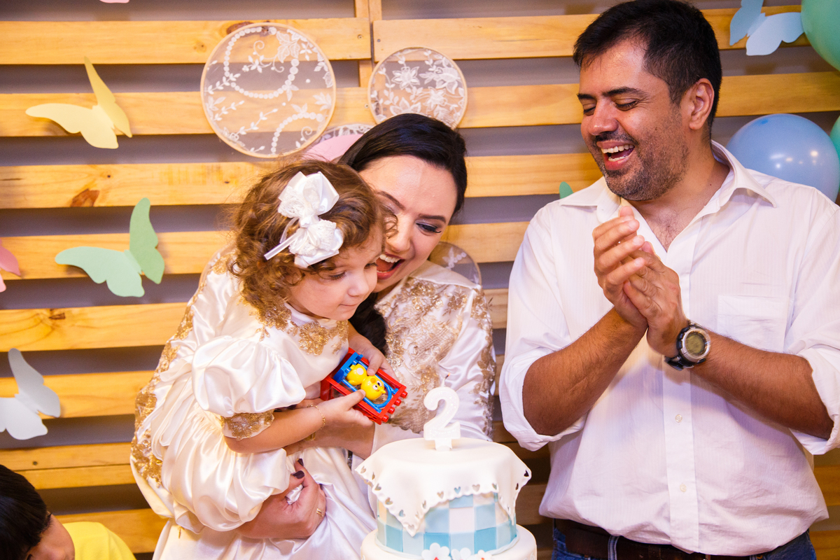 Criança no colo da mãe e ao lado do pai soprando as velinhas em seu aniversário de 2 anos.
