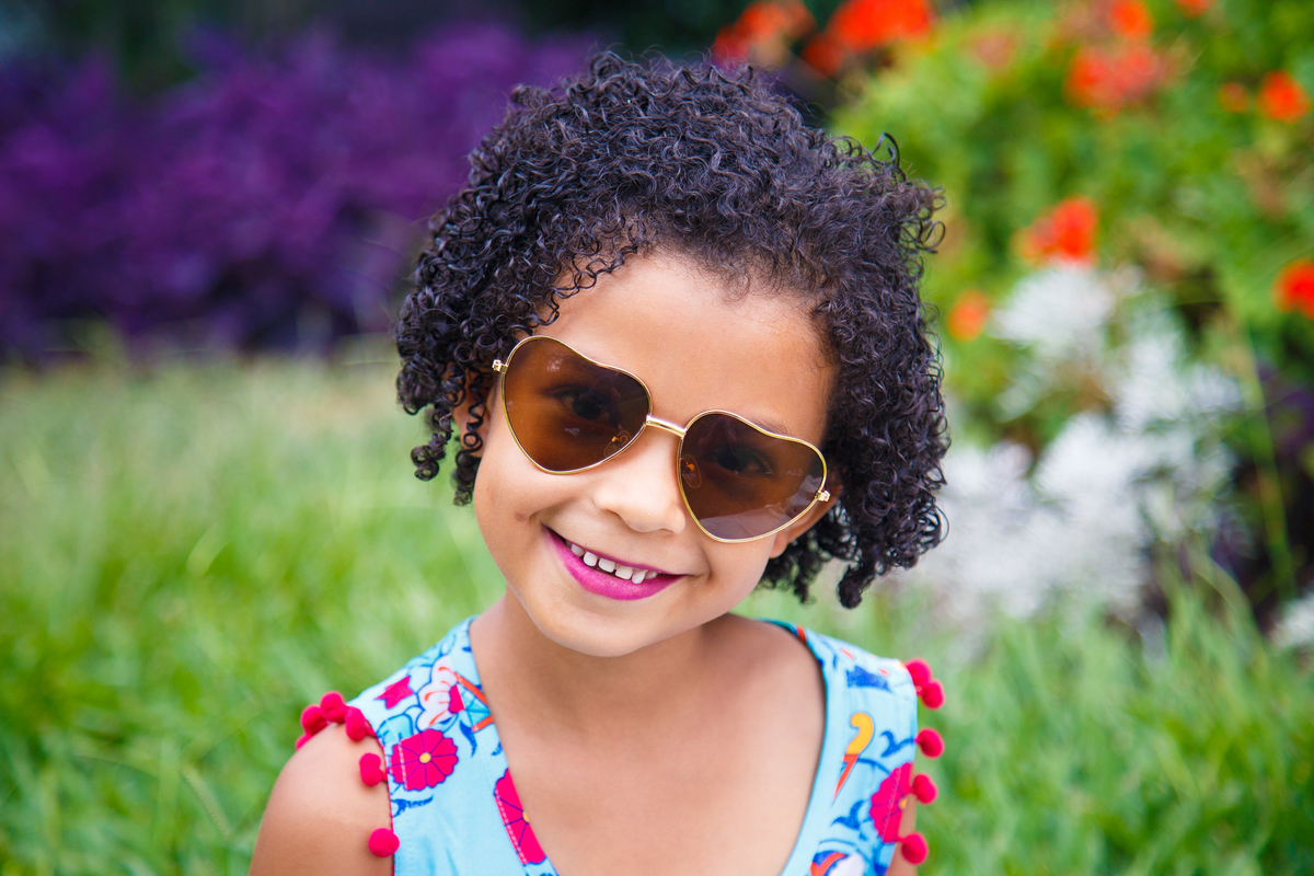 Criança usando um óculos de sol de coração, olhando para a câmera e sorrindo em um ensaio fotográfico em parque de BH.