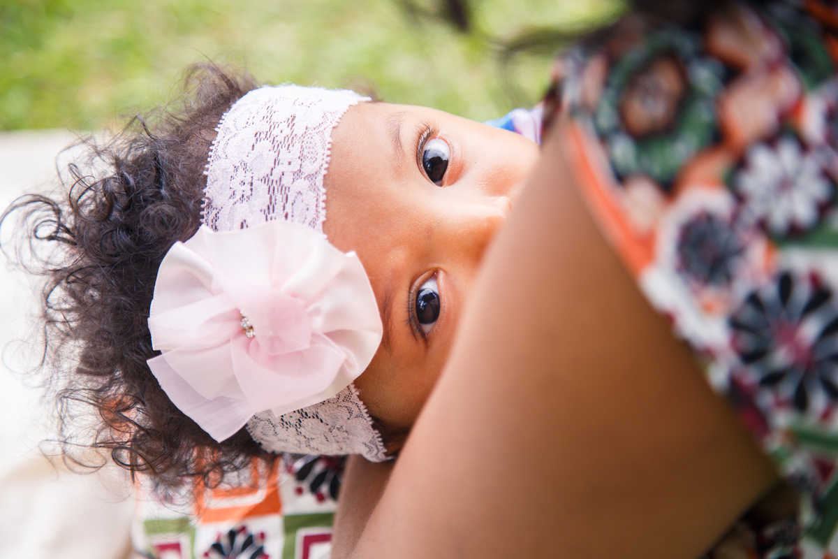Bebê mamando, a mãe de costas para a foto e o bebê de frente, aparecendo apenas dos olhos para cima, olhando para a câmera.
