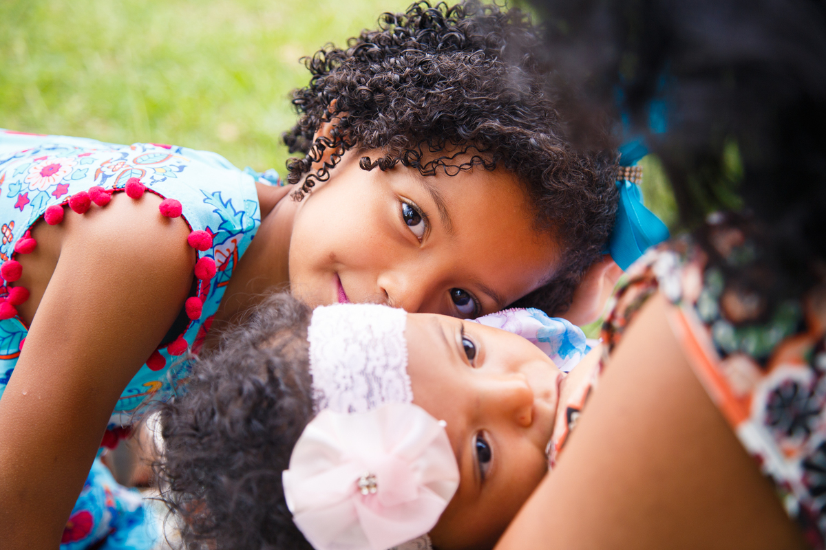 Bebê mamando e sua irmã dando um cheiro em sua cabeça.
