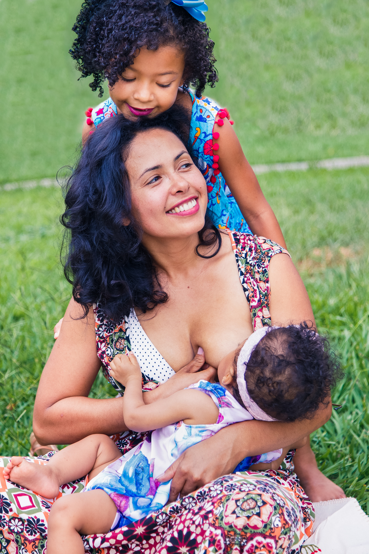 Mãe com suas duas filhas, uma mamando e a outra dando um abraço na mãe por trás.