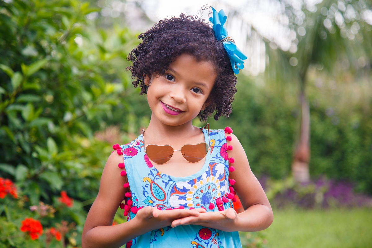 Criança posando para foto em um parque, olhando para a foto, sorrindo, com um óculos de sol pendurado no pescoço e as mãos abaixo do óculos.