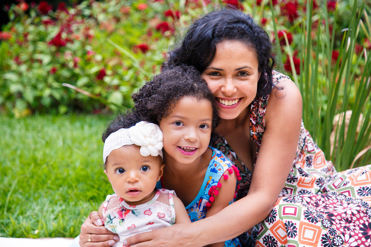 Mãe e suas duas filhas sentadas na grama, todas olhando para a câmera e se abraçando.