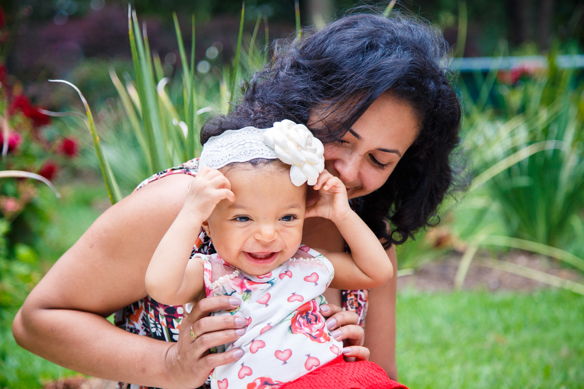Mãe e bebê brincando na grama. A mãe faz cócegas na bebê que sorri mexendo na tiara que está em sua cabeça.