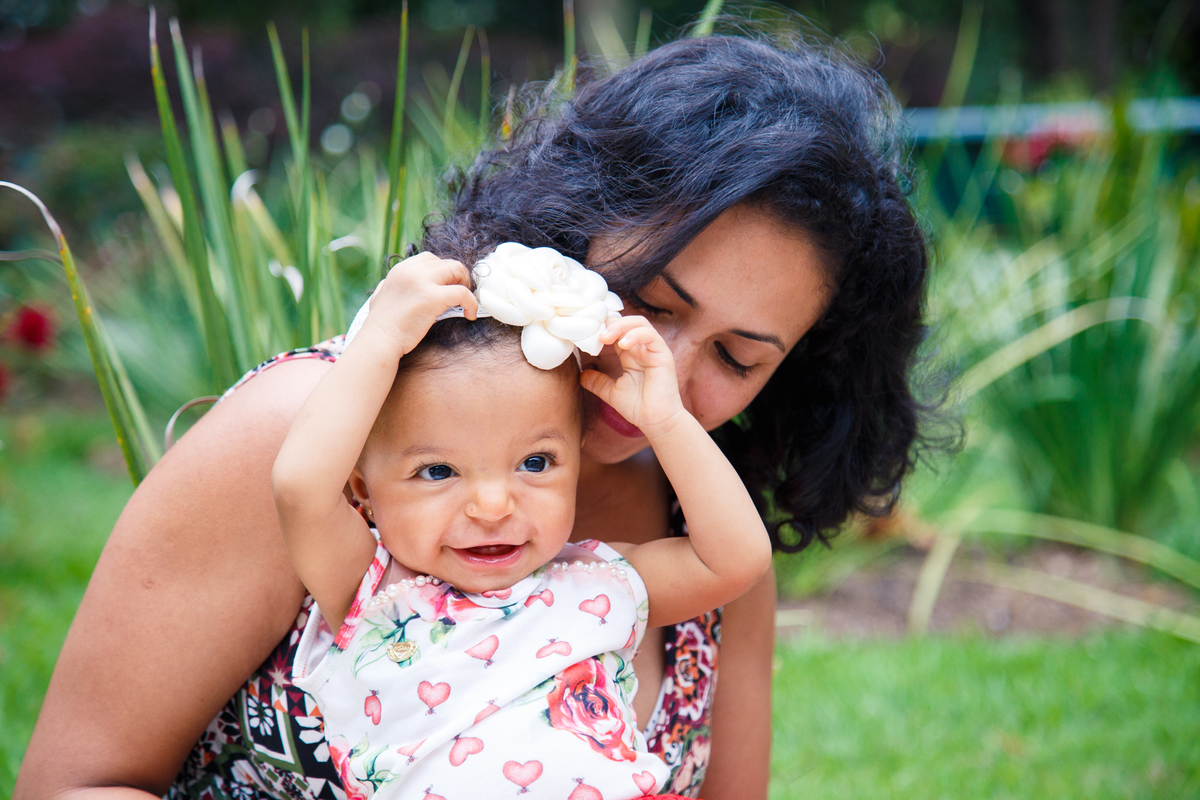 Mãe brincando com sua bebê sentada na grama. A mãe faz cócegas na bebê que ri e brinca com a tiara em sua cabeça.