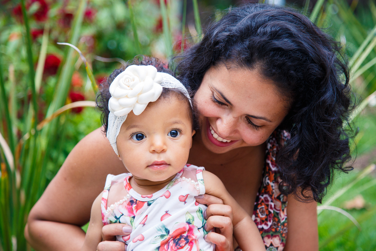 Mãe sentada na grama com sua bebê, a bebê olhando para frente e a mãe olhando a filha e sorrindo.