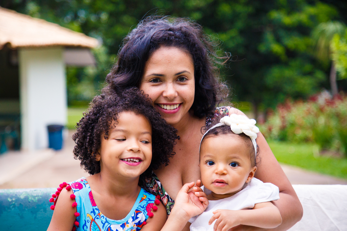 Mãe e as duas filhas sentadas em um banco. A mãe olha para a foto e a criança mais velha olha para a bebê e faz um carinho em sua bochecha.