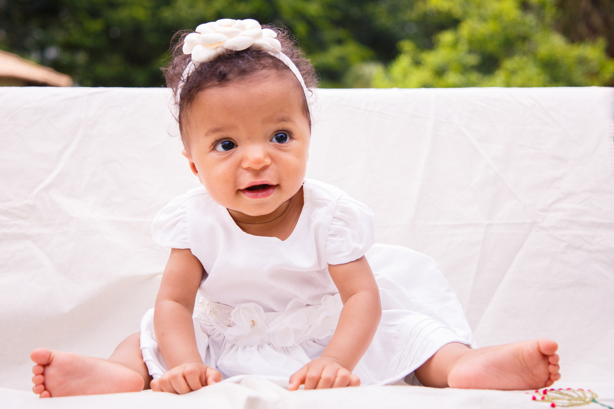 Bebê com roupinha de seu batizado sentada em um banco e sorrindo para a mãe que não aparece na foto.