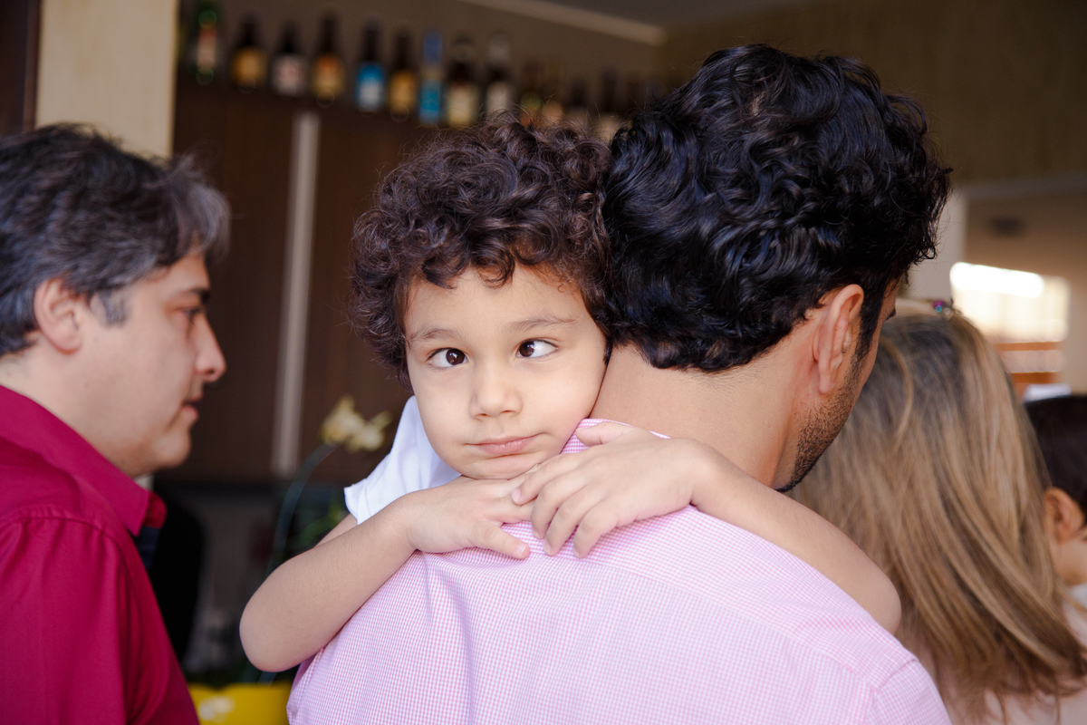 Criança no colo do pai, abraçando-o pelo pescoço e fazendo uma careta, ficando vesgo.