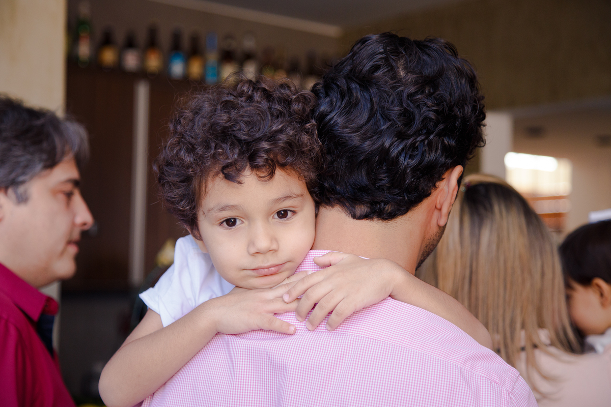 Criança abraçando seu pai pelo pescoço, o pai de costas e o rosto da criança em destaque na foto, com uma expressão singela e pensativa.
