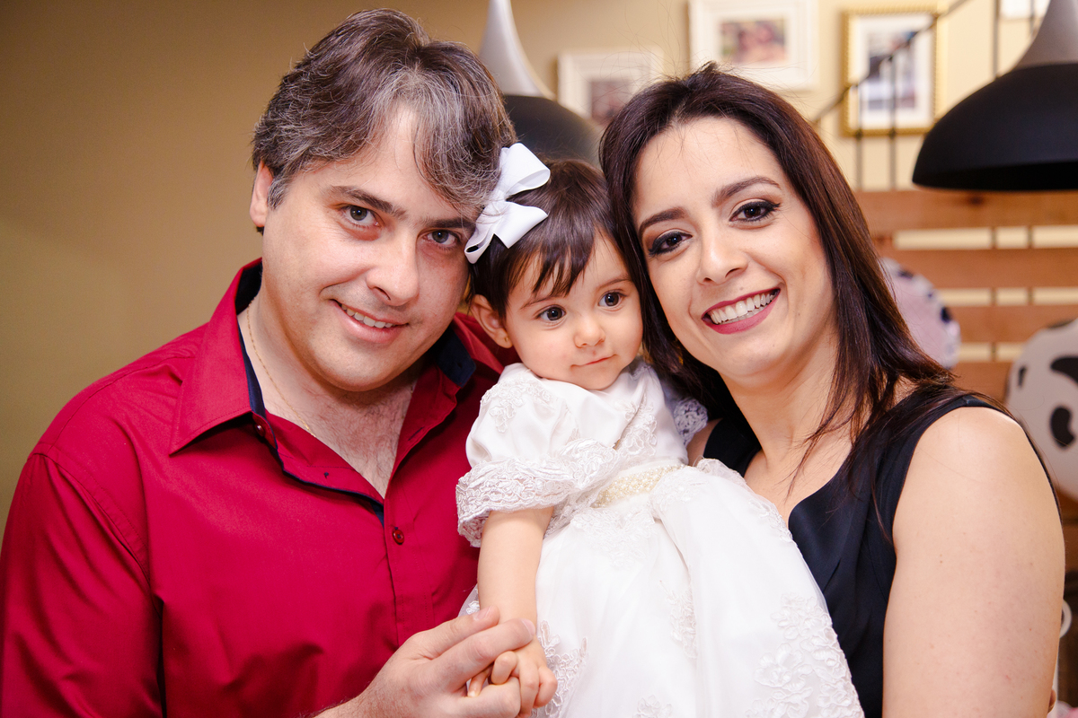 Bebê no colo de seu pai e sua mãe, os pais olham para a câmera e sorriem enquanto a bebê faz uma expressão de envergonhada.