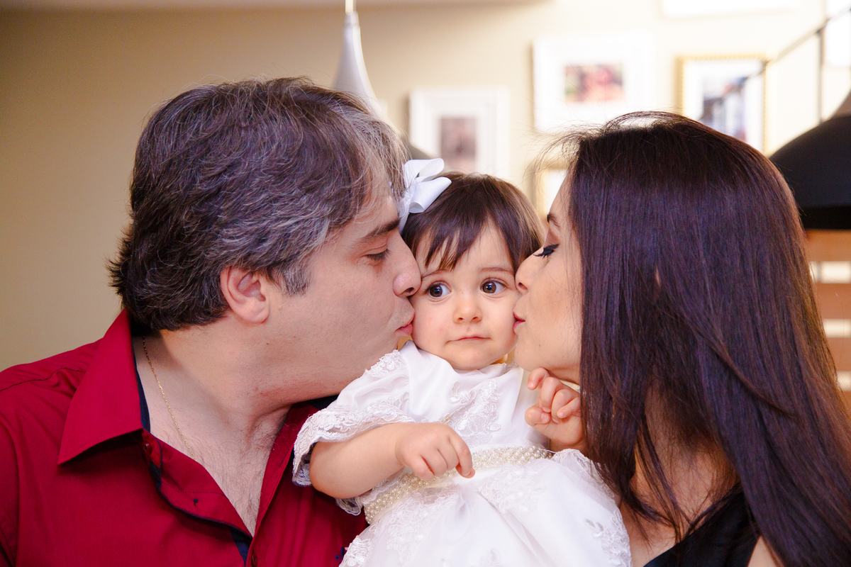 Bebê no colo de seus pais ganhando um beijo de cada um, em cada bochecha, em seu aniversário de 1 ano.