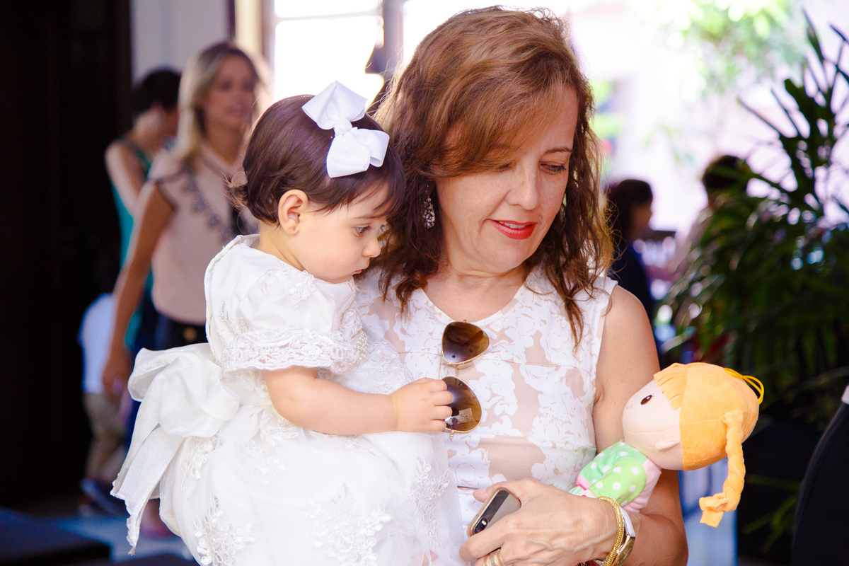 Bebê no colo da tia, que está segurando também uma boneca e ambas olham para o brinquedo.