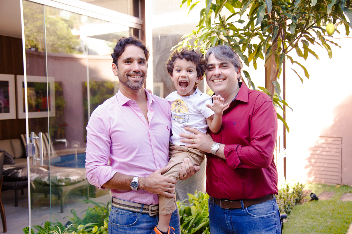 Criança no colo do pai e do tio fazendo uma careta para a foto enquanto os adultos sorriem.