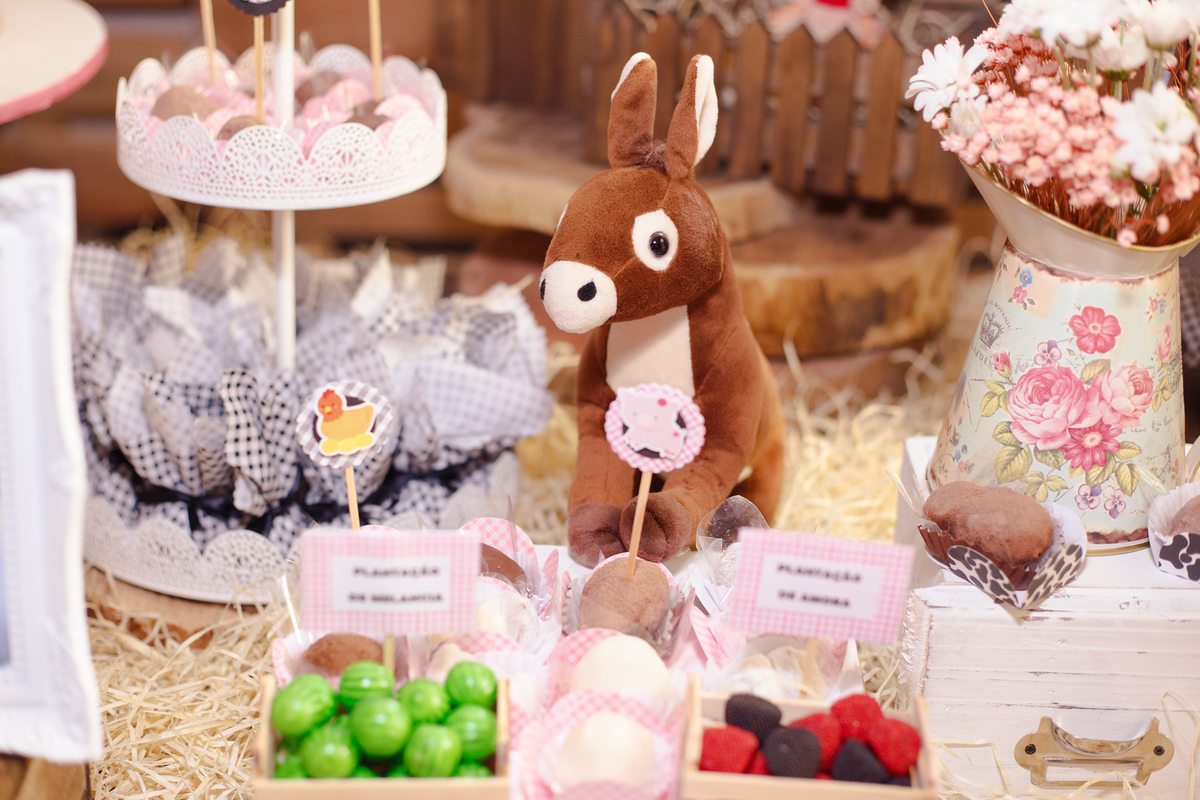 Detalhe de uma decoração de aniversário infantil no tema fazendinha, com um burro de pelúcia e algumas frutas em miniatura e um jarro com flores ao lado.