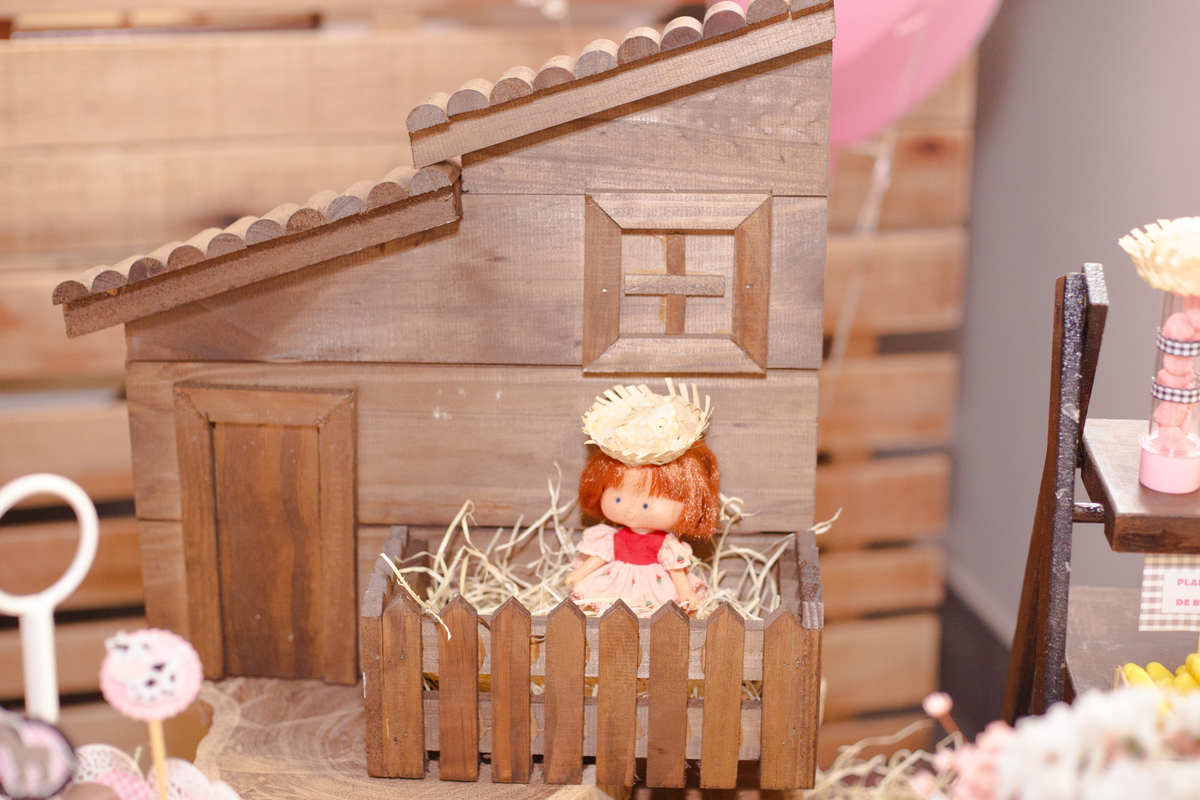 Detalhe da decoração de um aniversário infantil, sendo uma casinha de madeira com grade e uma boneca com chapéu de palha próxima à porta.