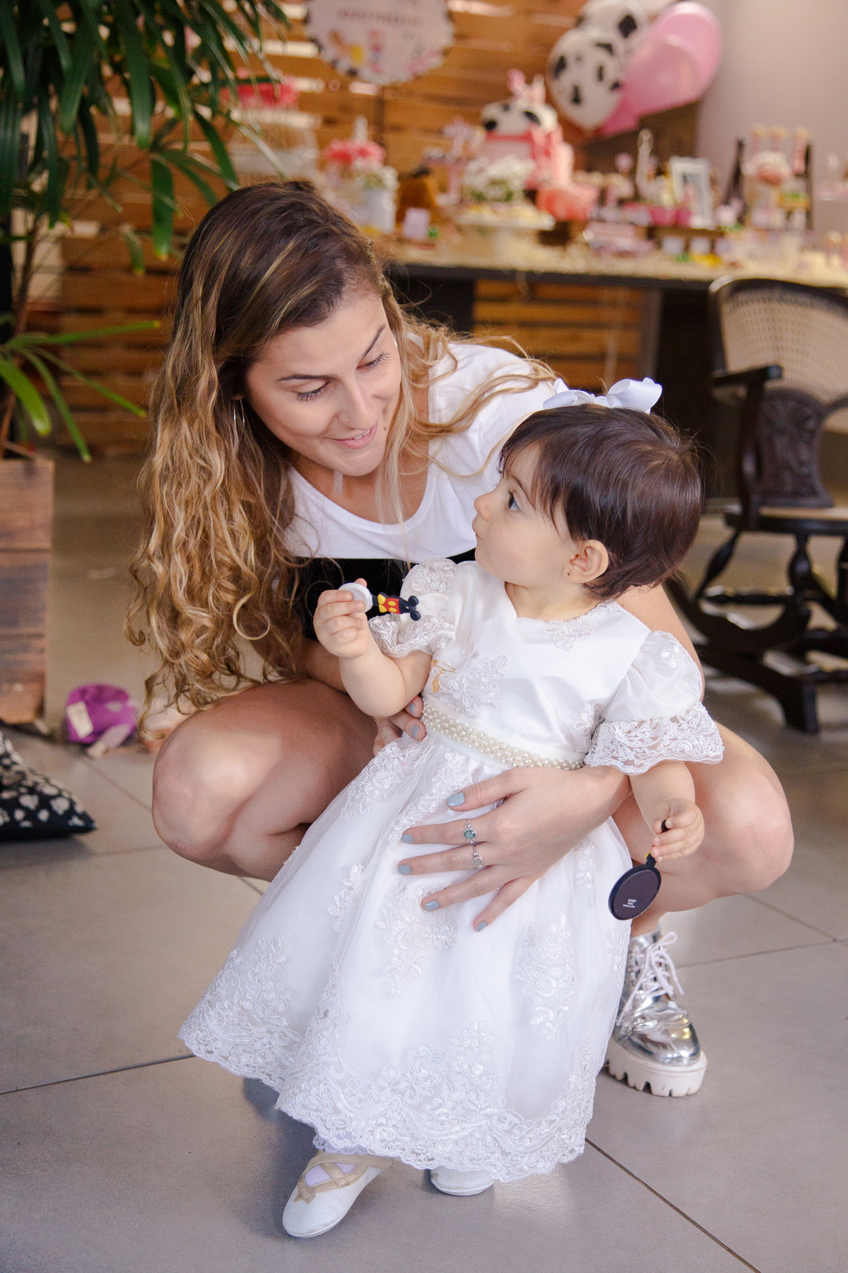 Bebê de pé no chão, sendo apoiada por sua prima, agachada ao seu lado, elas se olham e conversam.