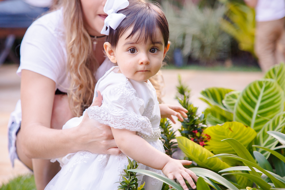 Bebê no jardim de sua casa, de pé, pegando em algumas plantas enquanto olha para a câmera.