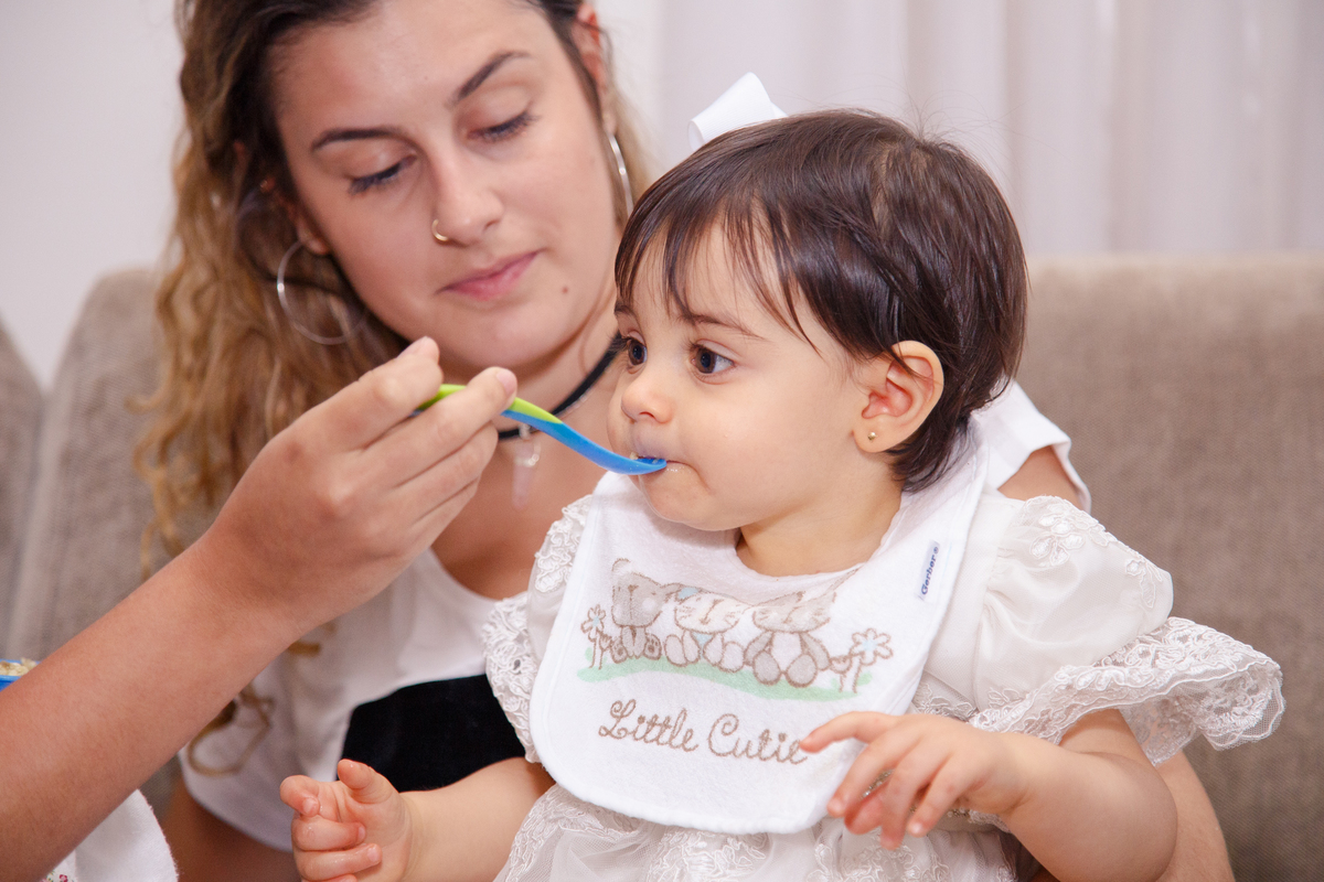 Bebê ganhando papinha em seu aniversário de 1 ano, ela está com babador e uma colher azul que a prima usa para levar comida à boca da bebê.