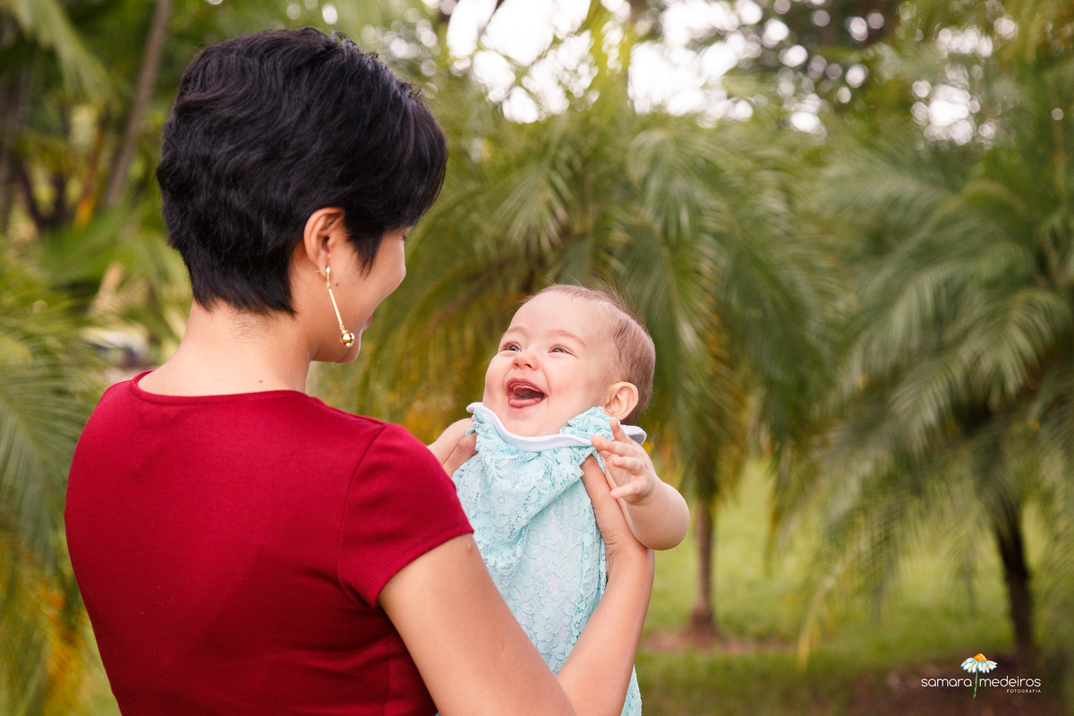 Criança no colo da mãe, que está de costas para a foto e o rosto da bebê em evidência com um sorriso enorme.