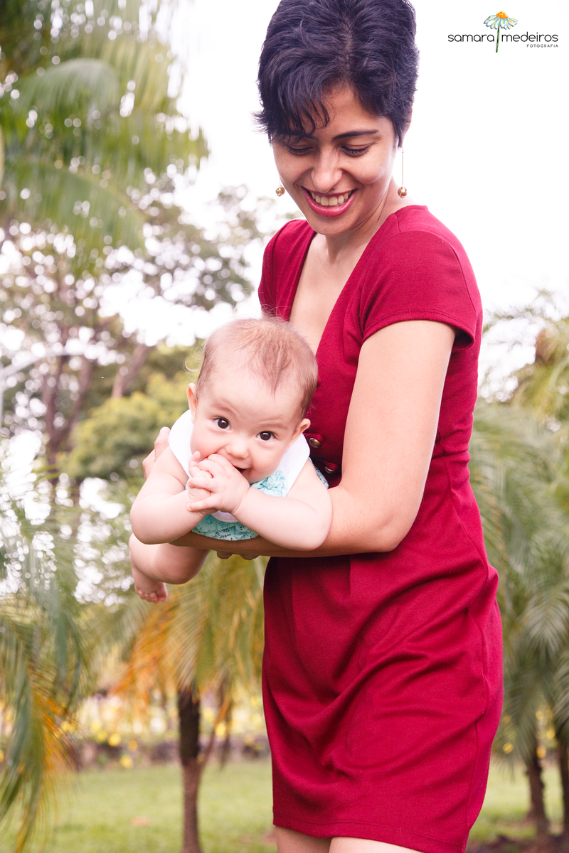 Mãe brincando de aviãozinho com a bebê, que está olhando para a câmera, sorrindo, com as mãozinhas na boca.
