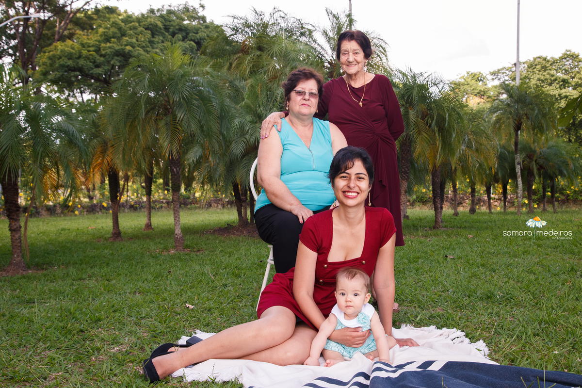Quatro mulheres em quatro gerações, posando para uma foto, mãe com a bebê sentadas no chão, a avó da bebê sentada em uma cadeira e a bisavó de pé ao lado.